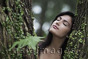 Asia Images Group - Head shot of young woman resting her head on a tree with eyes closed.
