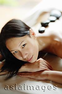 Asia Images Group - Woman lying on massage table, resting head on hands, stones along her back, looking at camera