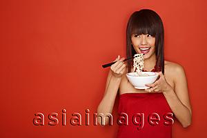 Asia Images Group - Young woman standing against red wall, eating a bowl of noodles