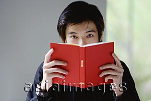 Asia Images Group - Young man with book covering his face, looking at camera