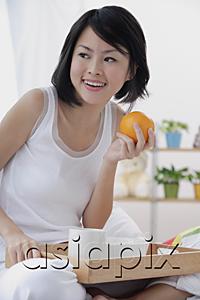 AsiaPix - Young woman having breakfast in bed, holding orange, looking away