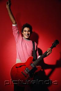 PictureIndia - Young man holding red guitar, standing against red wall, smiling at camera